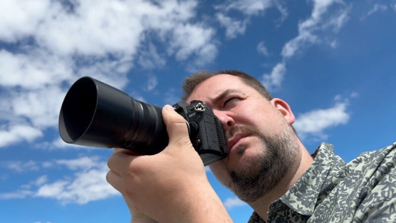 A man with a beard is looking through the viewfinder of a camera with a large lens, aiming upwards. He's wearing a patterned shirt, and a blue sky with scattered clouds is in the background.