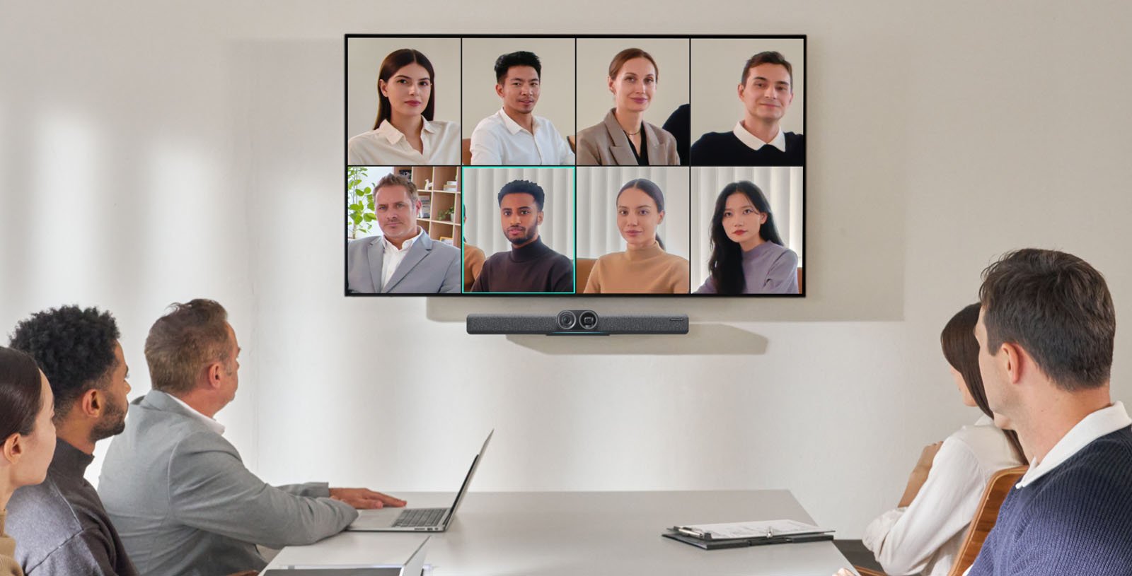 A group of people sitting at a conference table are engaged in a video call displayed on a large screen. The screen shows eight participants. A camera and speaker are positioned on the wall below the screen.