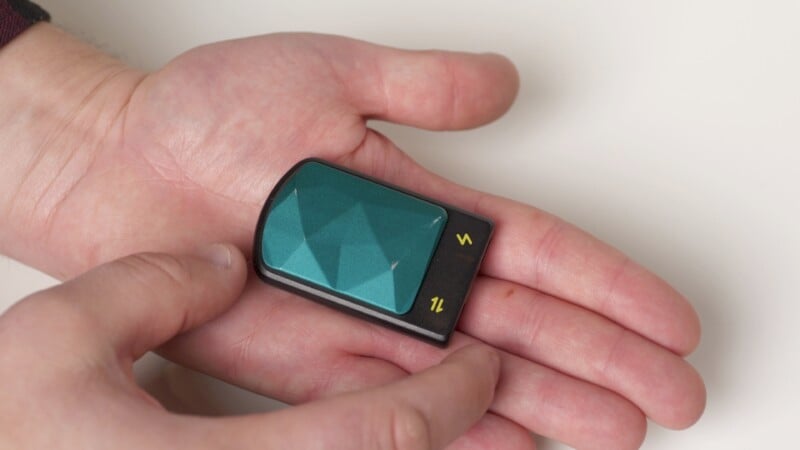 A person holds a small, teal, geometric-patterned electronic device with black edges in their palm. The device displays yellow symbols. The background is a plain, light-colored surface.