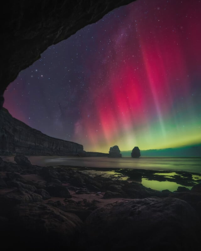 A stunning display of red and green auroras illuminates the night sky, casting a vibrant glow over a rocky beach. Two large rock formations are silhouetted against the colorful backdrop, creating a picturesque scene.