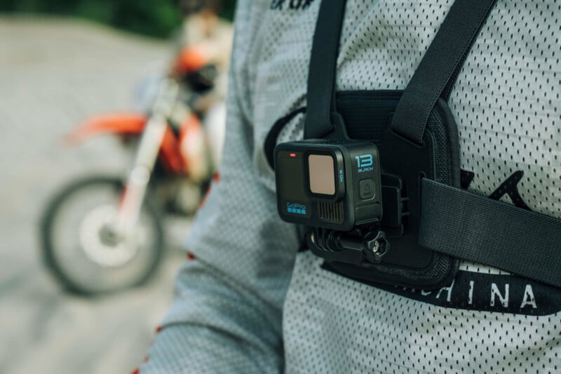 A person wearing a chest harness mounts a GoPro camera. A red motorcycle is blurred in the background. The focus is on the camera and harness setup.