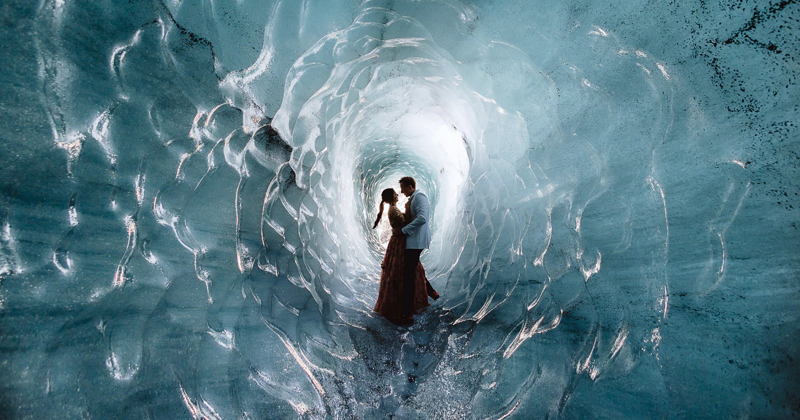 A couple stands in an embrace inside a stunning ice cave. The walls are smooth and glistening, illuminated by soft light filtering through the ice, creating an enchanting, otherworldly atmosphere. The couple appears silhouetted against the icy backdrop.