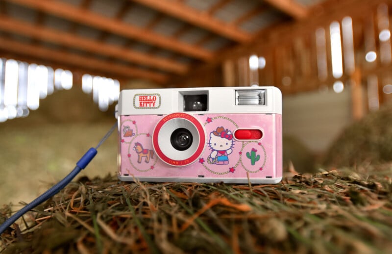 A pink Hello Kitty-themed camera with cartoon illustrations is resting on a pile of straw in a wooden barn. The camera has a blue wrist strap attached.