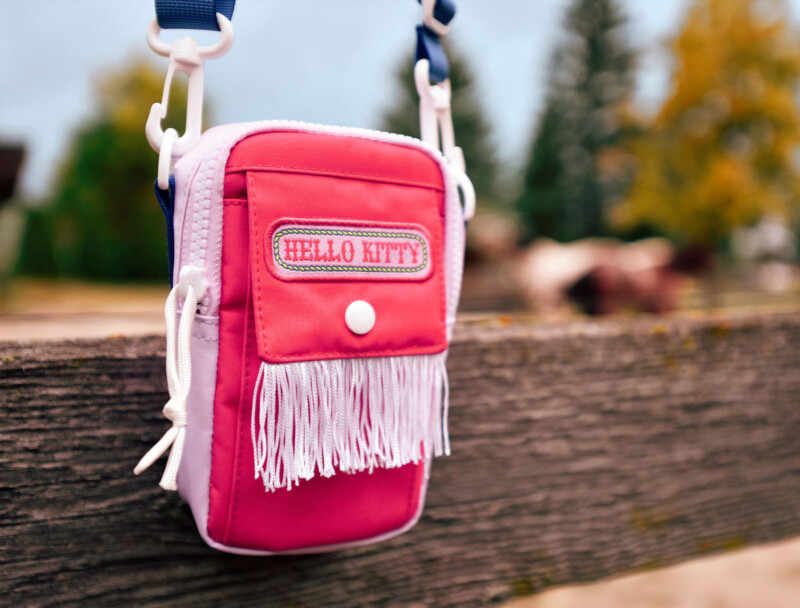 A small pink and red Hello Kitty crossbody bag with white fringe, hanging on a wooden fence. The background is slightly blurred, showing trees and a horse.