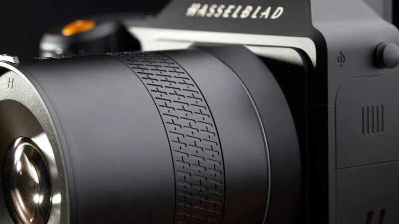 Close-up of a black Hasselblad camera, focusing on the textured zoom or focus ring of the lens. The brand name "Hasselblad" is visible at the top, with part of the lens and body shown in sharp detail against a dark background.