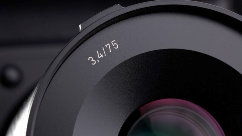 Close-up of a camera lens with an aperture marking of 3.4/75 on the rim. The lens is dark with a reflective surface showing a hint of color. The background is softly blurred.