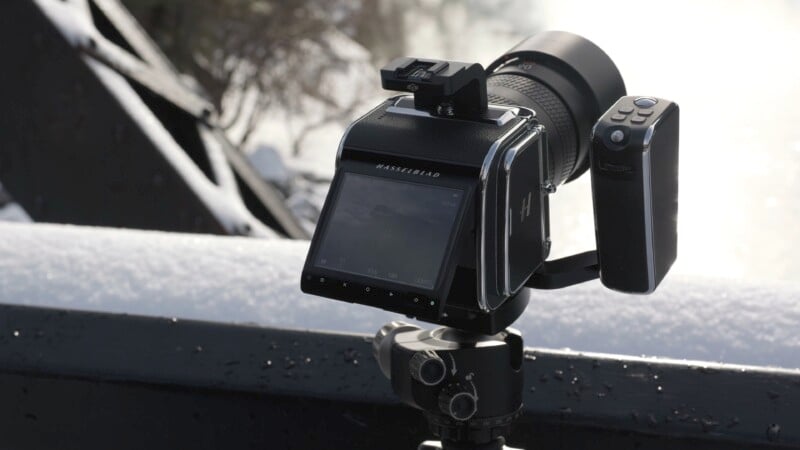 A professional Hasselblad camera is mounted on a tripod, positioned outdoors. The camera's screen is angled towards the viewer. The background features blurred natural elements and hints of sunlight reflecting on surfaces.