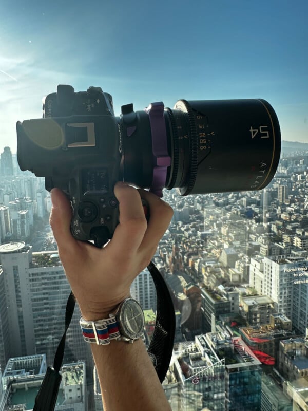 A person holds a camera with a large lens, pointing it at a cityscape from a high vantage point. The city below is bathed in sunlight, showcasing numerous buildings and a clear sky. The photographer's watch and part of the wrist strap are visible.