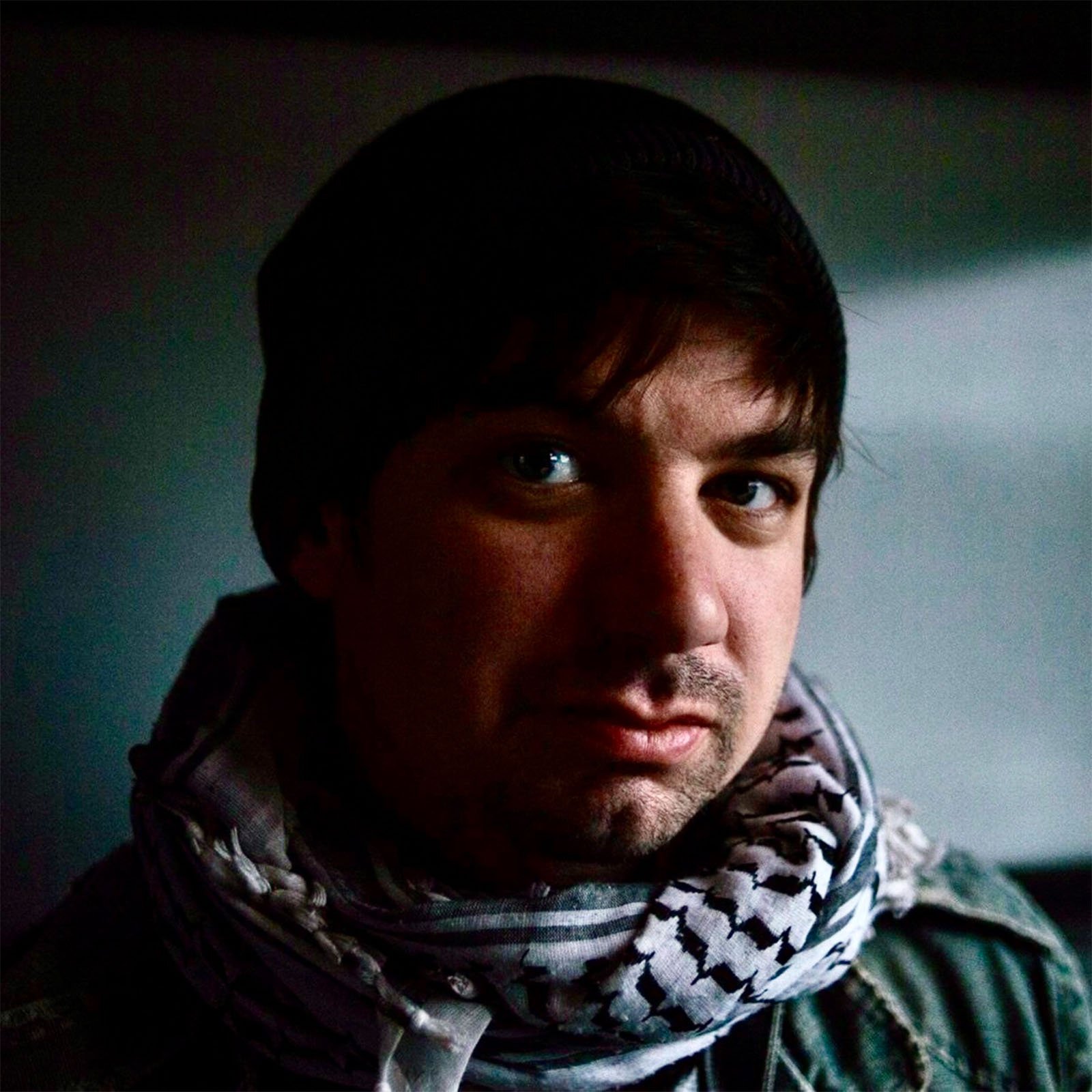 A person wearing a dark beanie, scarf, and jacket is looking at the camera. The background is dimly lit, casting a shadow over part of the face, highlighting a contemplative expression.