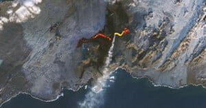 Aerial view of a volcanic eruption with bright orange lava flowing down a rocky landscape. Smoke and steam rise from the vent, and the dark terrain surrounding the lava contrasts with the blue ocean in the lower part of the image.