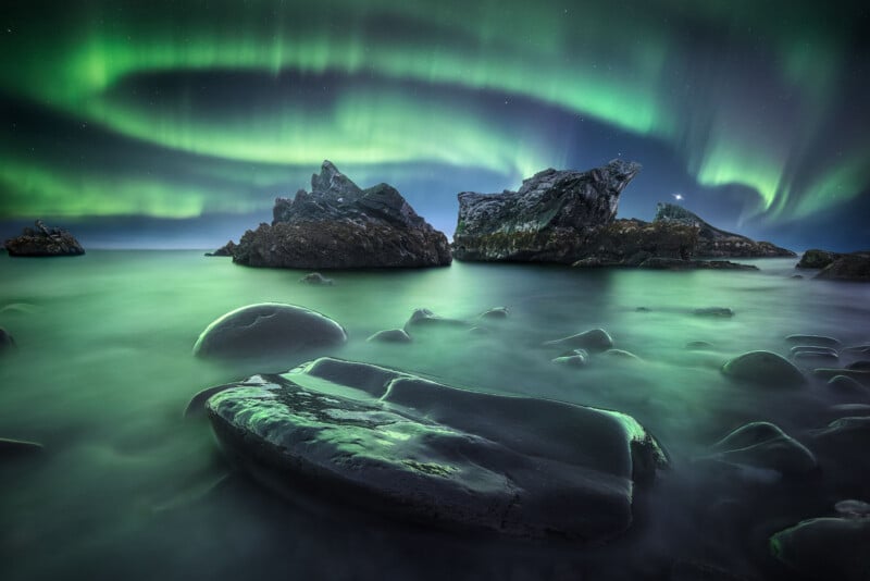 The image depicts a mesmerizing view of the Northern Lights over a rocky seashore. The green auroras create swirling patterns in the night sky, reflecting on the smooth water and rocks below.