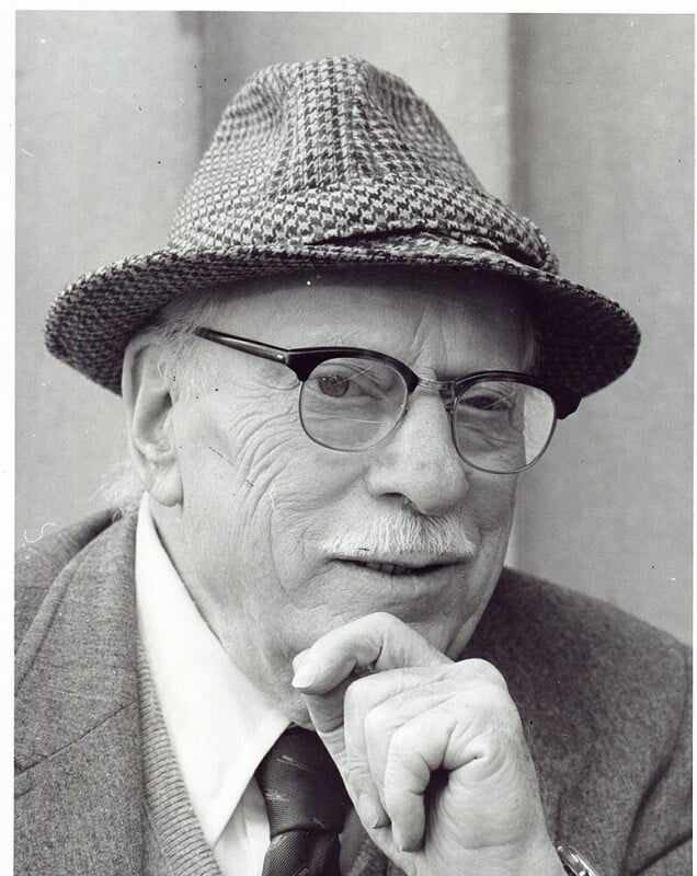 A black and white photo of an elderly man wearing glasses and a houndstooth-patterned hat. He has a thoughtful expression and is resting his chin on his hand. He is dressed in a suit jacket and tie.