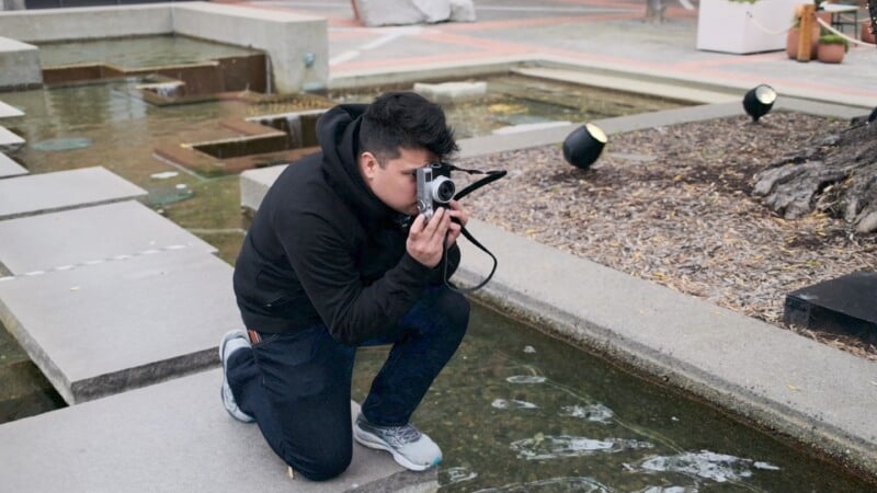 A person kneels by a shallow pond, holding a camera to their eye. They are wearing a black jacket and jeans. The scene includes concrete steps and lighting fixtures in an outdoor setting.