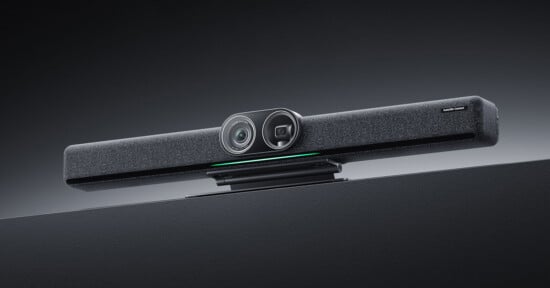 A sleek, modern video conferencing soundbar with dual cameras and a fabric-covered speaker, mounted on top of a large screen against a plain background.