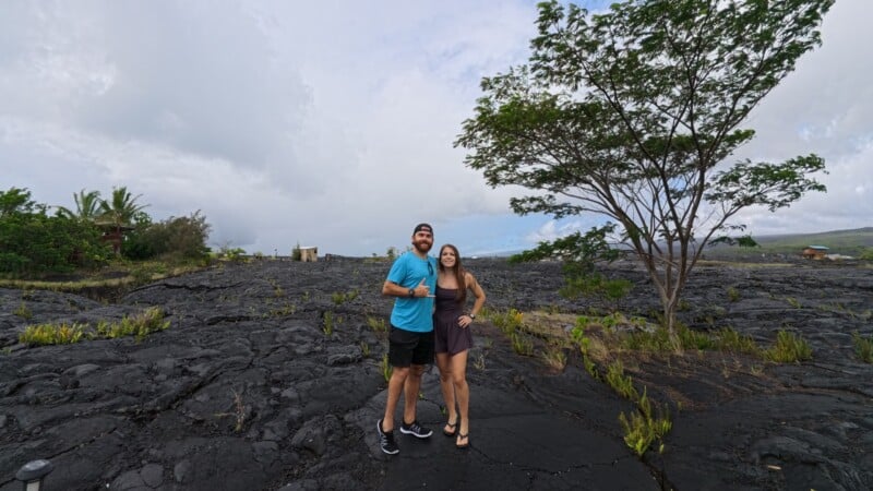 A smiling couple stands on a rocky black lava field under a cloudy sky. They pose arm in arm, with a tree on their right. Sparse vegetation and distant buildings are visible in the background.