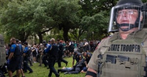 A state trooper in riot gear stands in the foreground, while a group of police officers and a crowd of protesters are in the background on a grassy area. Trees line the scene, creating a natural backdrop.