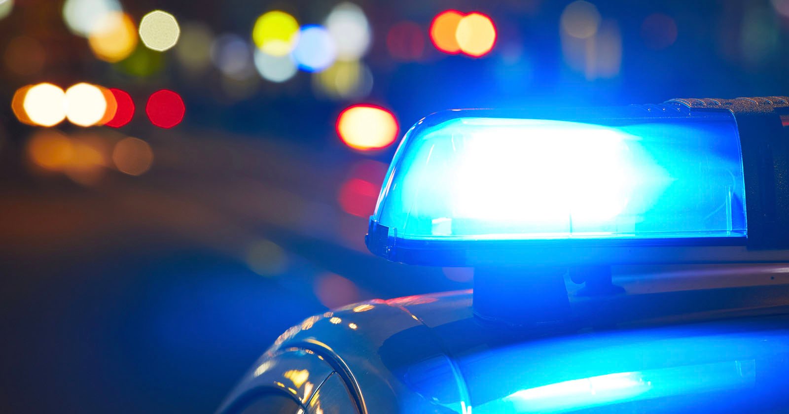 Close-up of a blue police car siren flashing brightly at night, with blurred colorful lights in the background, suggesting a busy urban street scene.