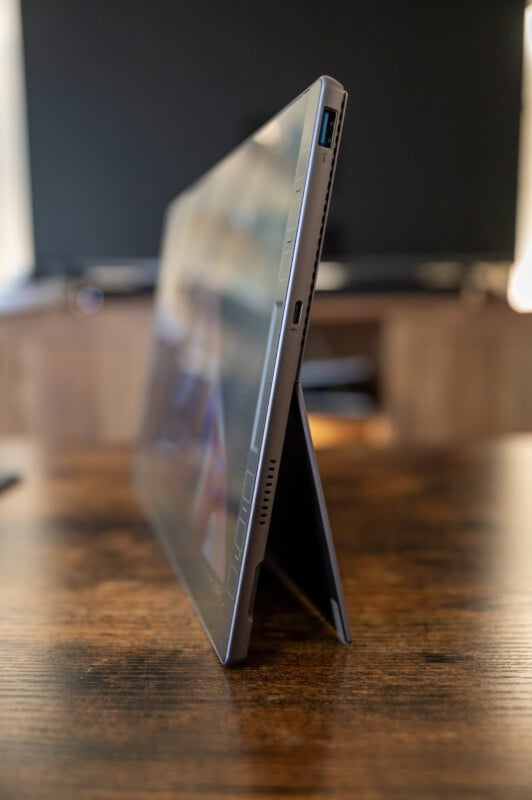 A sleek, thin tablet with a built-in kickstand is positioned on a wooden table. The side view displays several ports including a USB and microSD slot. The background is blurred, highlighting the tablet's modern design.