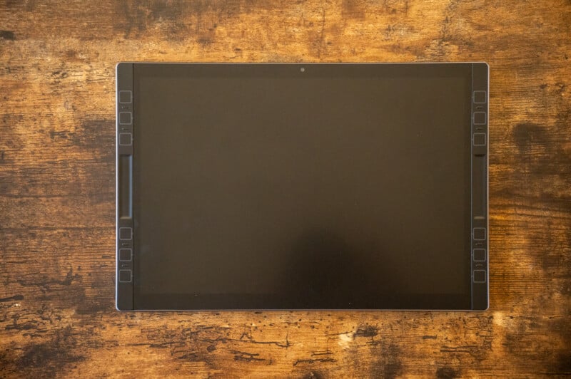 A digital drawing tablet with side buttons is placed on a textured wooden surface. The screen is turned off, reflecting a faint outline of a person's head and camera.