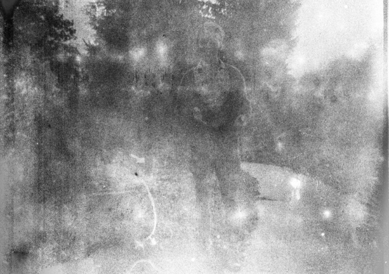 A grainy, black and white image with a silhouetted figure standing outside. The background is blurred, with indistinct trees and sky. The overall effect is atmospheric and abstract.