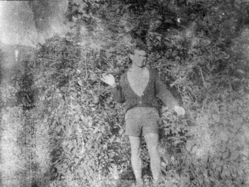 A grainy, black-and-white photograph of a person standing in front of dense foliage. The person is wearing a light-colored shirt, a dark jacket, and shorts. They are holding one arm out slightly to the side. The image has a vintage feel.