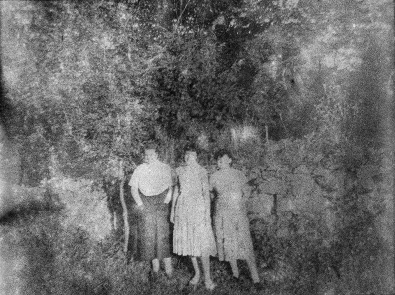 A black and white photo of three people standing outdoors. They are in front of a stone wall and surrounded by dense foliage, possibly trees and bushes. The image has a grainy, vintage appearance.