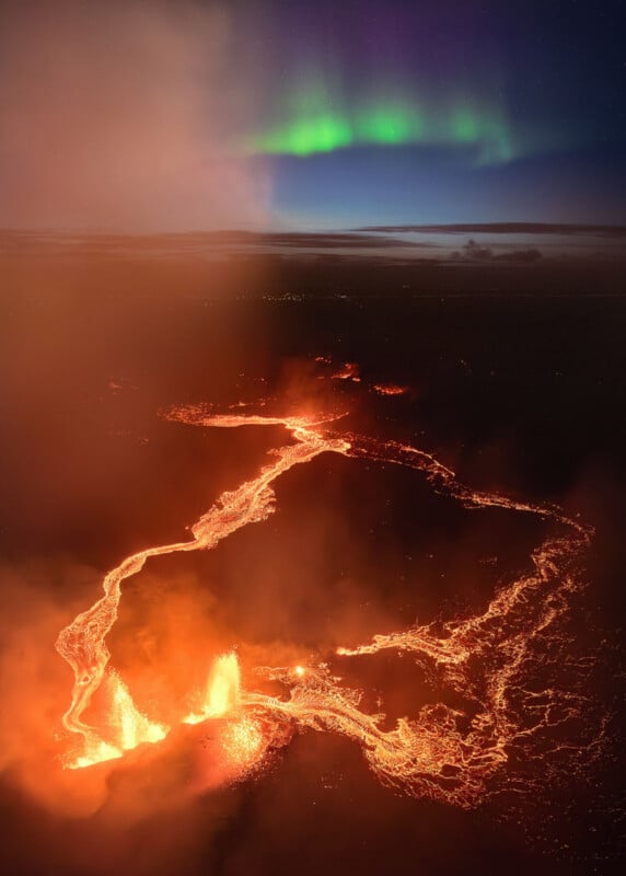 A spectacular landscape shows a glowing lava flow and eruption in the foreground with vibrant, swirling northern lights in the night sky above. The contrast of fiery orange and cool green creates a stunning visual display.