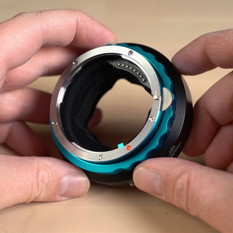 A person holding a camera lens adapter with a blue and black metallic finish. The adapter features an electronic contact point and a circular opening. The person's fingers are visible, gripping the sides of the adapter on a light wood surface.