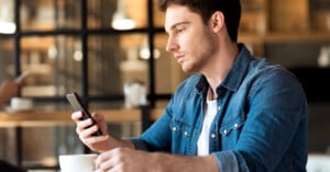 A man in a denim shirt sits at a wooden table in a café, looking at his smartphone. His right hand holds a cup, and the background features blurred shelves and another person using a phone. The scene is softly lit and relaxed.