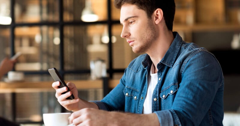 A man in a denim shirt sits at a wooden table in a café, looking at his smartphone. His right hand holds a cup, and the background features blurred shelves and another person using a phone. The scene is softly lit and relaxed.