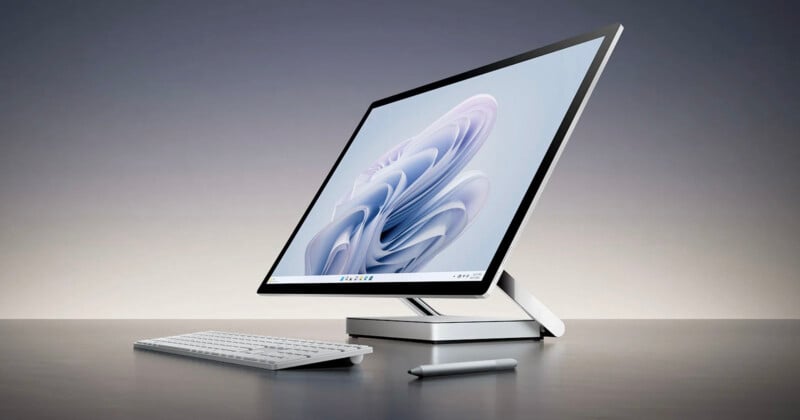 A sleek, modern desktop computer with a large screen displays an abstract design. It is accompanied by a slim keyboard and a stylus, all set on a smooth gray surface against a gradient background.