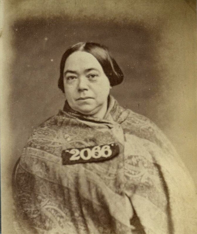 A sepia-toned image of a woman with dark hair parted in the middle, wearing a patterned shawl. She is facing the camera and appears to have a serious expression. She is wearing a tag with the number "2068.