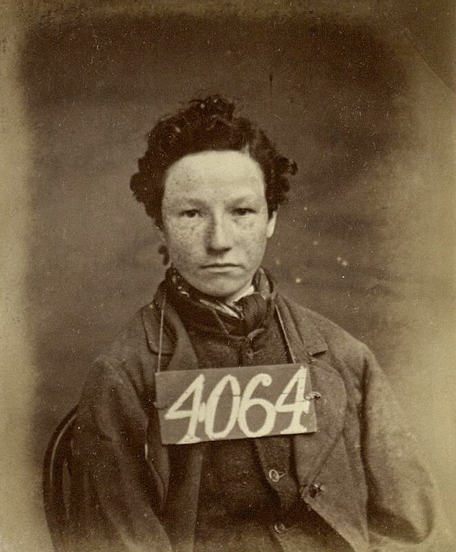 Sepia-toned vintage portrait of a young person with curly hair, wearing a suit and a number placard "4064" hanging from the neck. The background is plain.