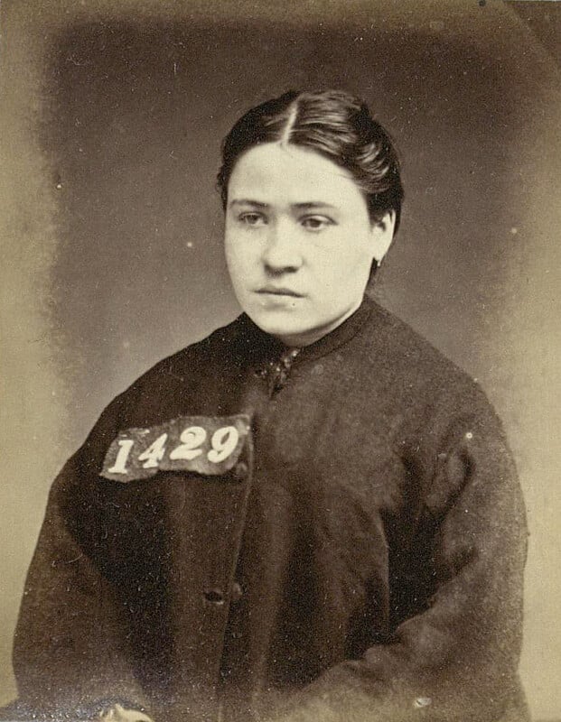 A vintage black-and-white photograph of a woman with dark hair, dressed in a plain dark garment. She is wearing a badge with the number "1429" displayed on it. The background is a solid color, giving the image a classic, historical look.