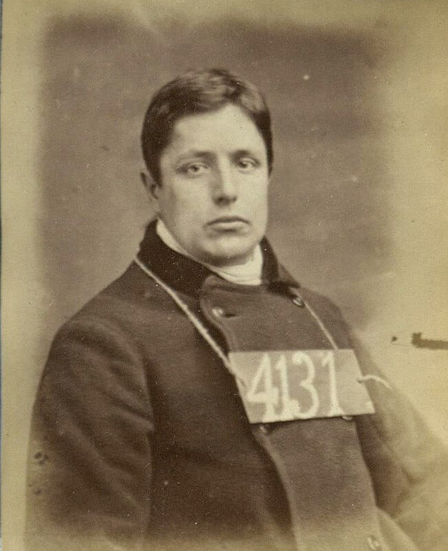 A black and white vintage photograph of a person wearing a dark coat. The person has a neutral expression and is facing the camera. A sign with the number "4137" is hanging around their neck. The background is plain and blurry.