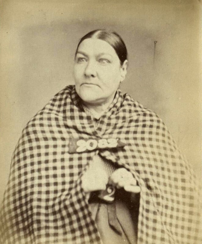 A vintage portrait of a woman with her hair pulled back, wearing a checkered shawl and holding a number card "2087." The image is sepia-toned, giving it an old-fashioned appearance.