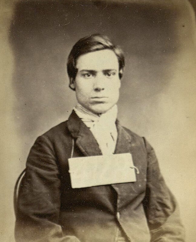 A black-and-white vintage photograph of a young man with short hair, wearing a dark jacket and a white collared shirt. He has a blank rectangular card attached to the front of his jacket. The background is plain and blurred.