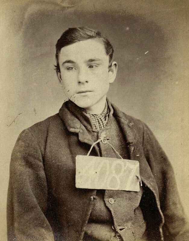 Sepia-toned vintage photo of a young boy wearing a dark jacket and a plaid shirt. He has short hair and gazes slightly to the side. A rectangular tag with the number "187" is hanging around his neck.