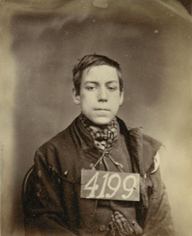 A young person with a serious expression sits facing forward, holding a card with the number 4199. They wear a worn jacket and scarf. The image is sepia-toned, with a vintage appearance.