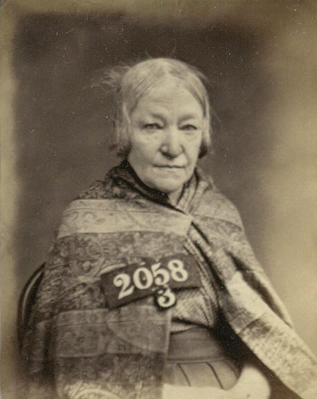 An elderly woman with a serious expression sits, wearing a patterned shawl. A placard with the number "2058" is displayed in front of her. The image is in sepia tone, giving it an aged appearance.