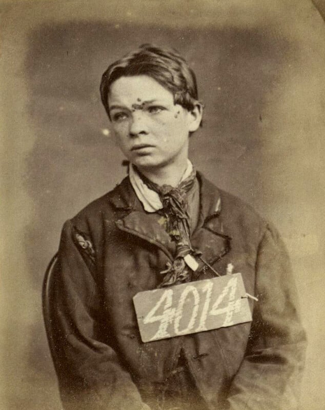 A black and white vintage photograph of a young person with short hair, wearing a jacket and a scarf. They have a faded sign with the number "4014" around their neck. The image has an aged, historical appearance.