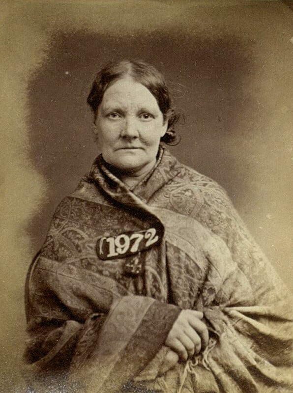 A sepia-toned vintage photograph shows a woman with dark hair pulled back, wearing a patterned shawl. She holds a placard with the number "1972." The background is plain, highlighting the woman's expression and attire.