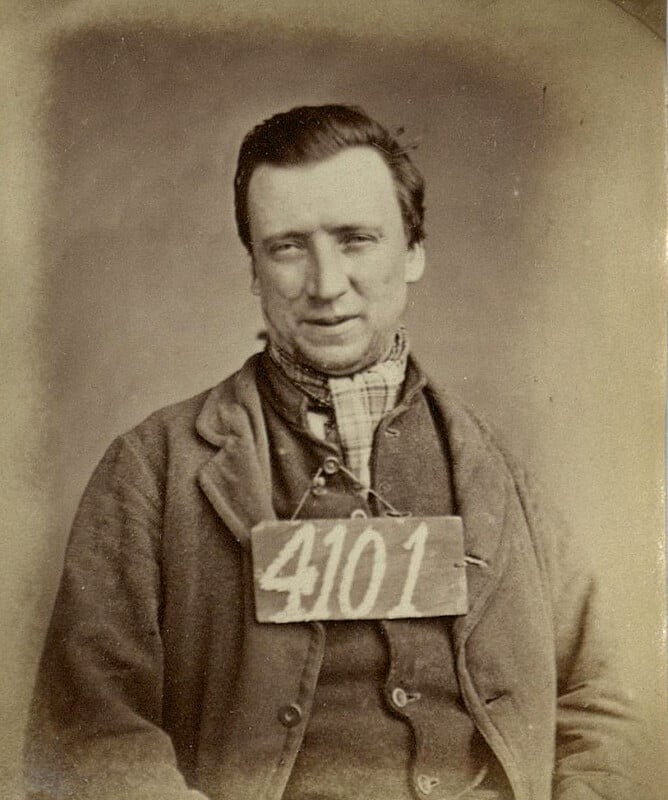 Sepia-toned portrait of a man with short hair, wearing a suit and scarf. He has a tag labeled "4101" hanging around his neck. The image has a vintage, historical look.