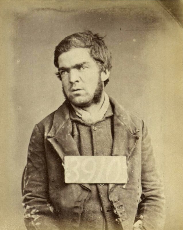 A sepia-toned photograph of a man with mutton chop sideburns, wearing a worn-out jacket and holding a placard with the number 3911. He has a serious expression and looks slightly to the left.