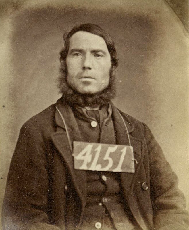 Sepia-toned image of a man from an older era, wearing a coat and sporting mutton chops. He has a paper with the number "4151" attached to his chest. The background is plain.