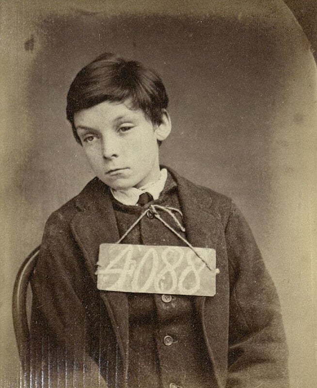 A vintage black and white photograph of a young boy with tousled dark hair wearing a suit and tie. He is seated with a numbered sign, "4088," hung around his neck. The expression on his face is neutral.