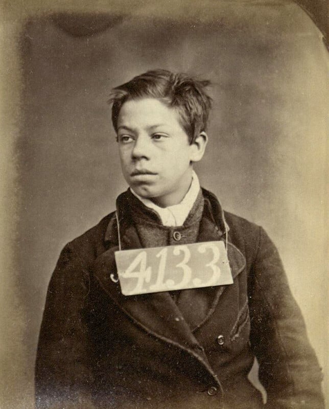 A vintage sepia-toned photograph of a young boy wearing a dark coat. He has short, tousled hair and is looking to the side. A rectangular card with the number "4133" is hanging around his neck. The background is plain and neutral.