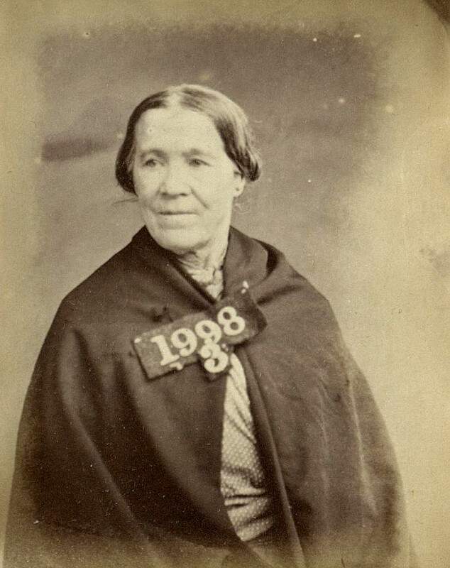 Black and white photo of an older woman with her hair tied back, wearing a dark shawl and a patterned gown. She has a tag with the number "1938" on her chest. The background is plain and neutral.