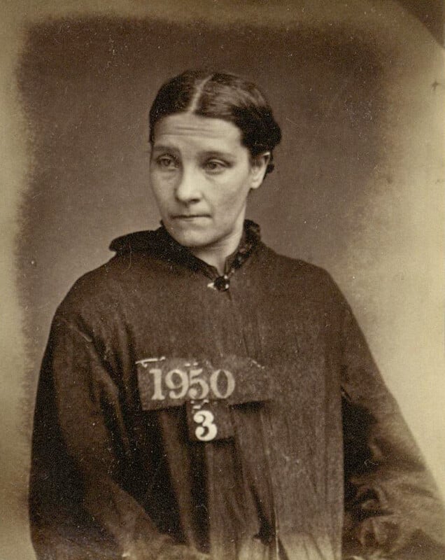 A sepia-toned photograph of a woman with dark, pulled-back hair, wearing a buttoned, dark garment. She has a somber expression and wears a sign with the numbers "1950" and "3" on her chest.