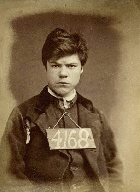 A sepia-toned photograph of a young person with short hair, wearing a dark jacket and a tag with the number 4168 hanging from their neck. The expression is serious, and the background is plain.